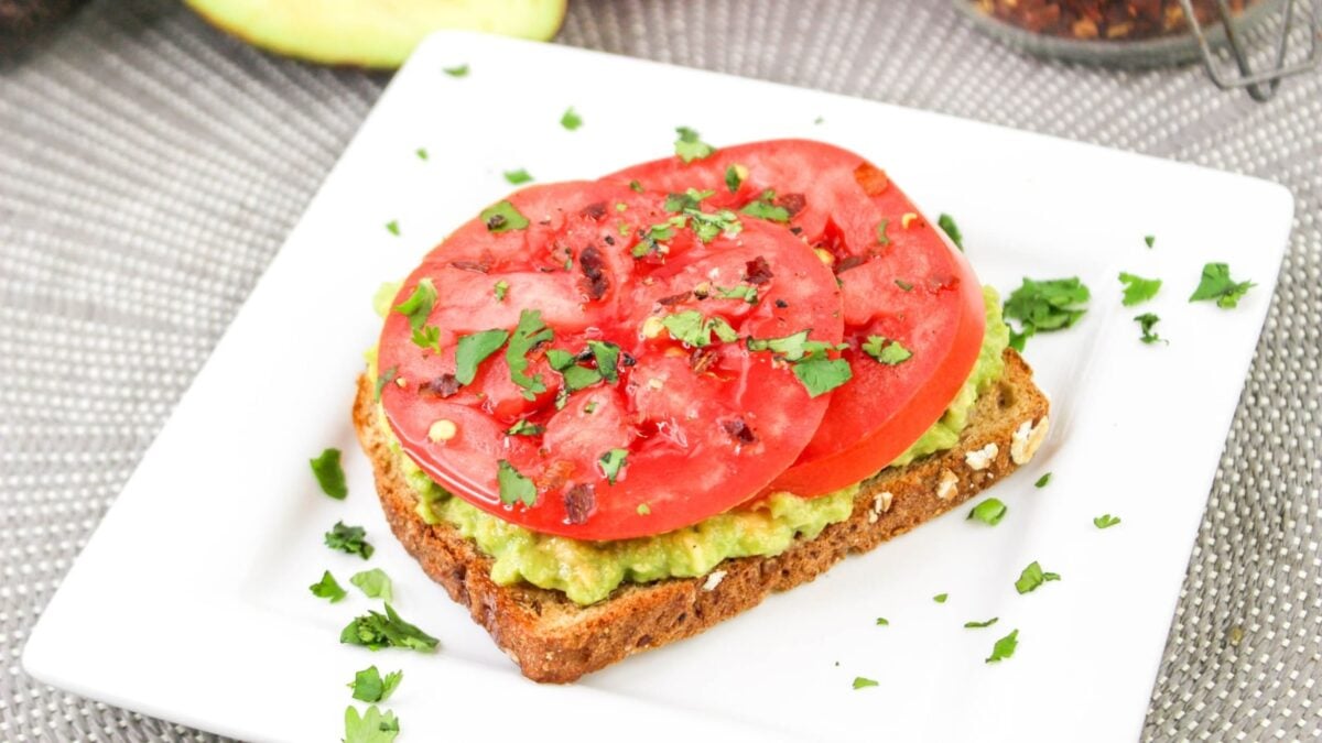 A slice of whole grain bread topped with mashed avocado, tomato slices, chopped cilantro, and red pepper flakes on a white plate—perfect for easy breakfast recipes or quick weekend breakfasts.