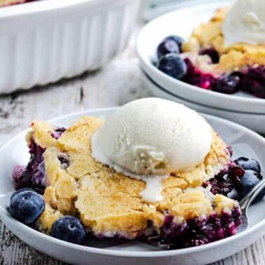 A serving of blueberry cobbler topped with a scoop of vanilla ice cream is on a white plate with a spoon, with fresh blueberries visible—one of the best desserts for fans of classic dessert recipes.