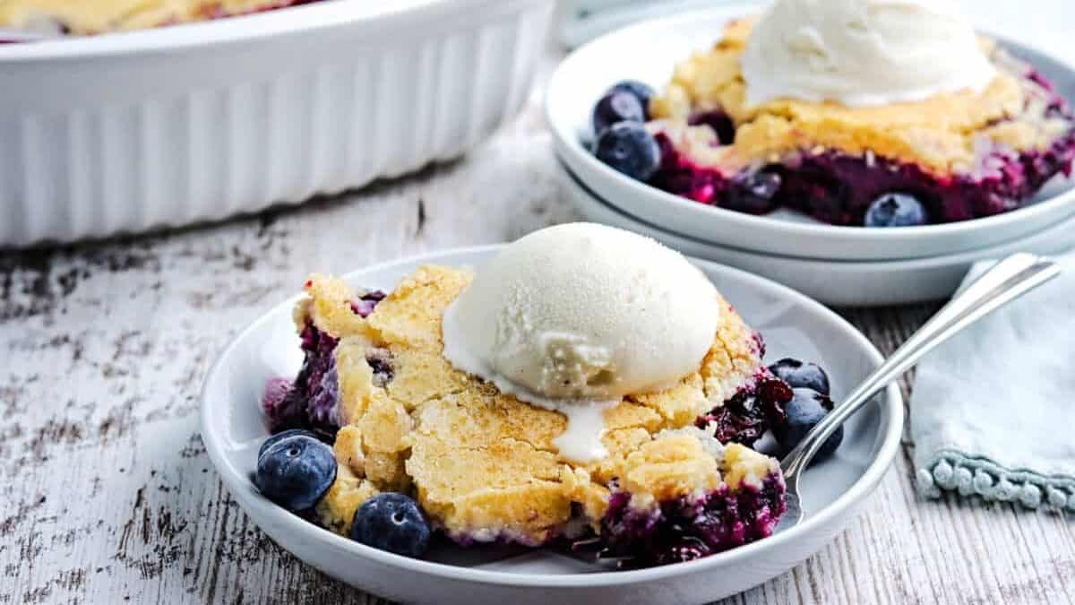A serving of blueberry cobbler topped with a scoop of vanilla ice cream is on a white plate with a spoon, with fresh blueberries visible—one of the best desserts for fans of classic dessert recipes.
