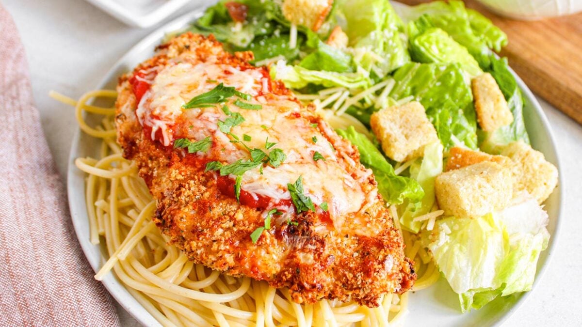 A plate with breaded chicken topped with tomato sauce and cheese on spaghetti, served with a Caesar salad and croutons—a true family favorite among Parmesan mains.