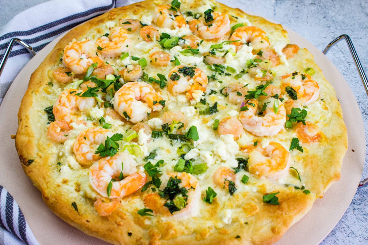 A baked unconventional pizza topped with shrimp, crumbled cheese, chopped herbs, and a white sauce, displayed on a round pizza stone with a striped towel nearby.