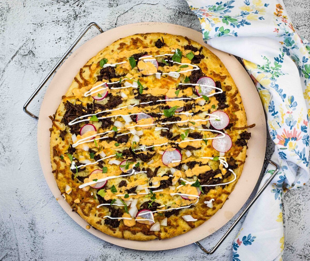 A unique pizza topped with ground meat, cheese, sliced radishes, onions, herbs, and drizzled with white sauce, served on a round baking tray next to a floral-patterned cloth.
