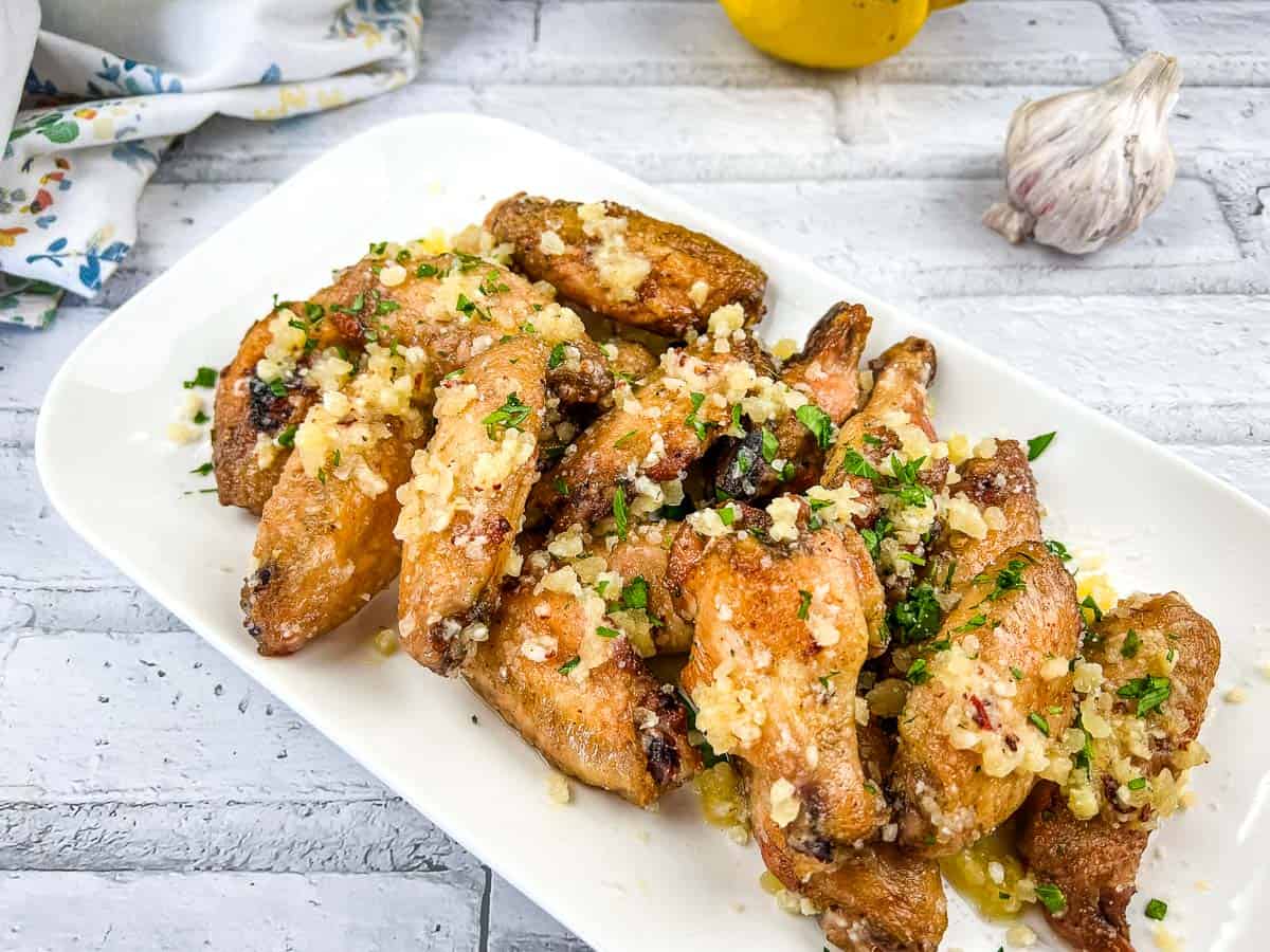 A white rectangular plate with fried chicken wings topped with minced garlic and herbs, set on a white brick surface with a garlic bulb and lemon in the background—perfect for adding to your list of Family Favorites or exploring new Parmesan Recipes.