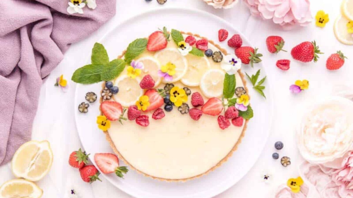 A lemon tart on a white plate, decorated with strawberries, blueberries, sliced lemons, mint leaves, and edible flowers, surrounded by fruit and flowers—perfect for those who love old fashioned dessert recipes and traditional desserts.