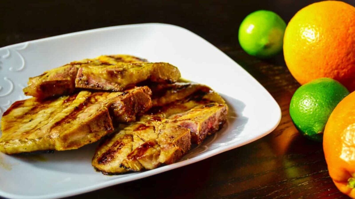 Grilled pork chops are stacked on a white plate, with whole oranges and limes placed nearby on a dark surface—perfect inspiration for your next BBQ dinners.