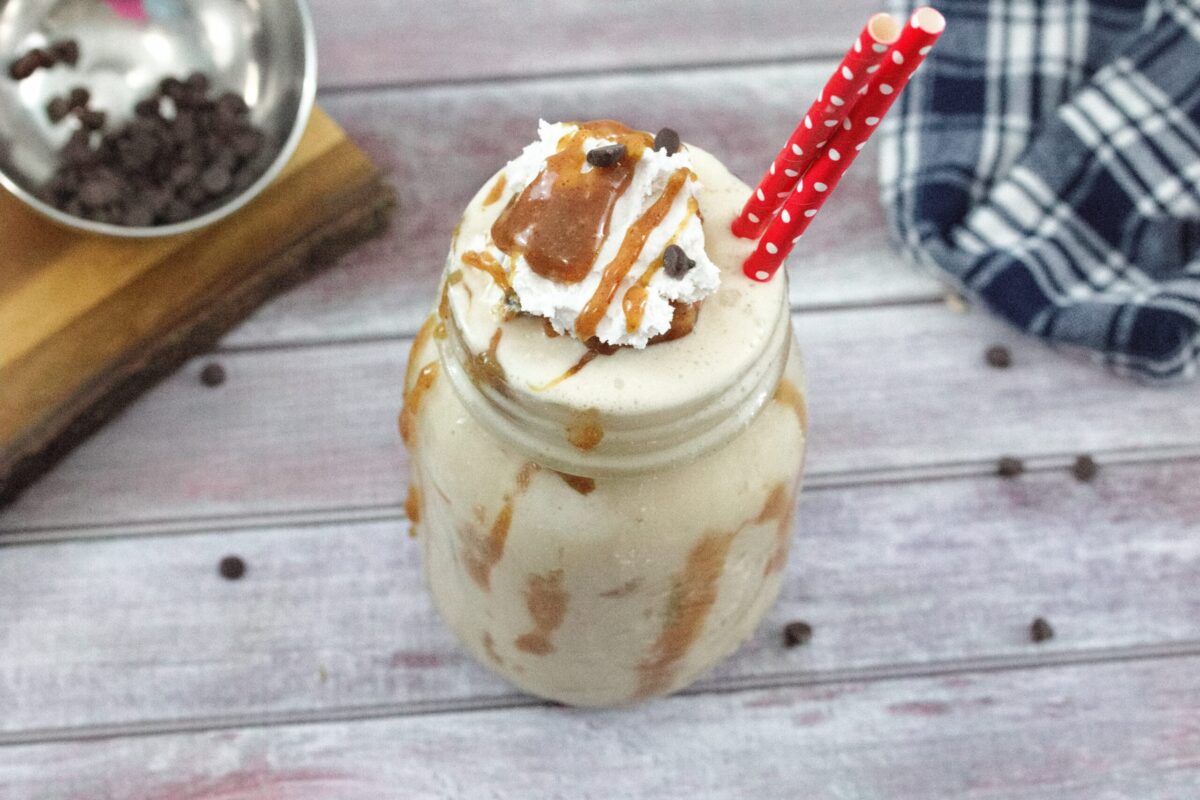 A mason jar filled with a creamy milkshake topped with whipped cream, caramel sauce, chocolate chips, and two red polka-dot straws makes one of the most tempting easy snacks on a wooden table.