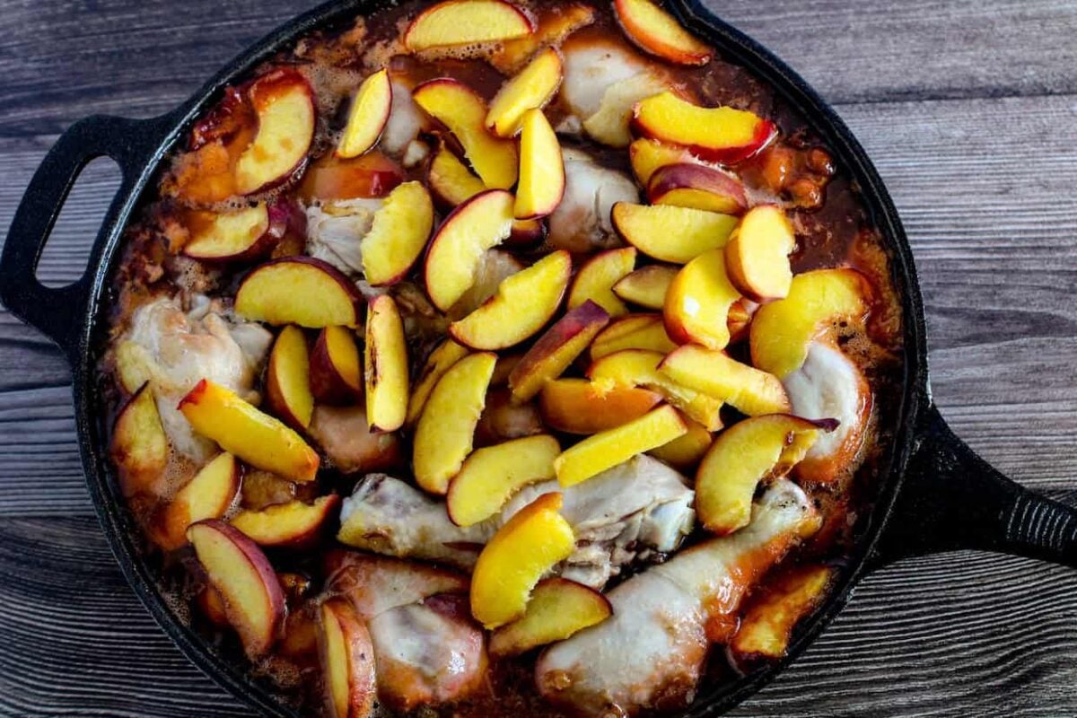 A cast iron skillet filled with cooked chicken drumsticks, sliced fresh peaches, and a reddish-brown sauce makes for mouthwatering BBQ dinners on a wooden surface.