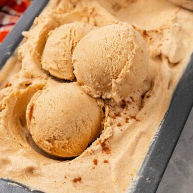 Three scoops of light brown frozen dessert in a rectangular metal container, with a sprinkle of spice on top and a checkered cloth in the background—a true Frozen Foods treat that's dangerous after midnight.