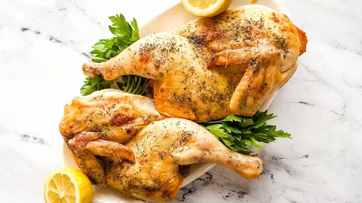 Two roasted Cornish hens on a white platter, garnished with fresh parsley and lemon halves, make an elegant roast dinner and a true showstopper recipe, beautifully presented on a marble surface.