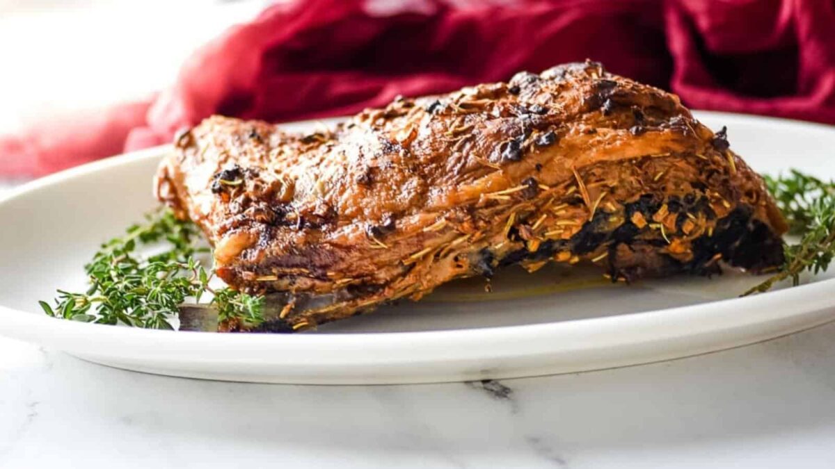A cooked, seasoned lamb shoulder rests on a white plate, garnished with sprigs of fresh herbs—a perfect centerpiece for fans of roasted dishes—set against a blurred red cloth background.