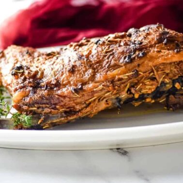 A cooked, seasoned lamb shoulder rests on a white plate, garnished with sprigs of fresh herbs—a perfect centerpiece for fans of roasted dishes—set against a blurred red cloth background.