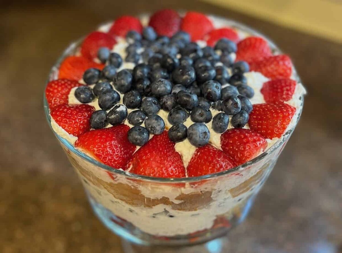 A glass trifle dish filled with layers of cake, cream, strawberries, and topped with blueberries and halved strawberries—perfect for those who love fruit dessert recipes or crave refreshing summer desserts.