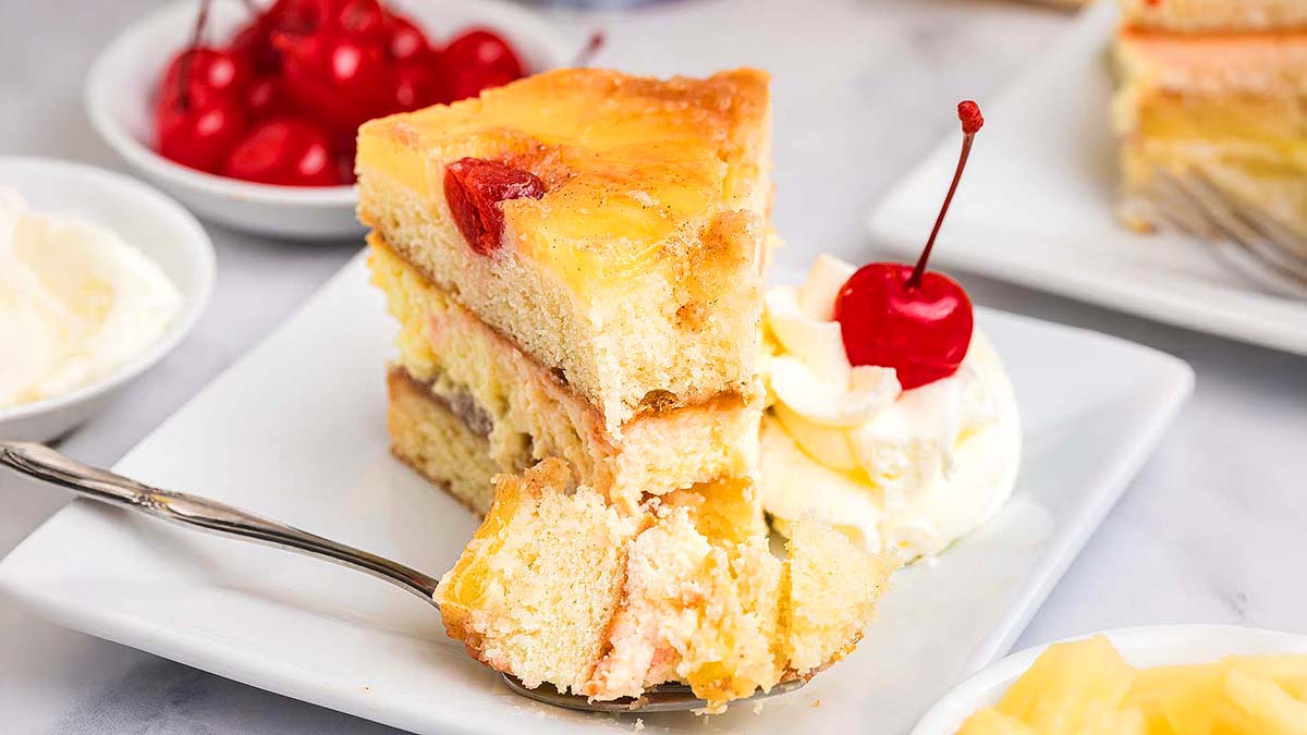 A slice of layered pineapple upside-down cake with whipped cream and a cherry on a white plate, perfect for lovers of fruit desserts, with a fork and a bowl of cherries in the background.