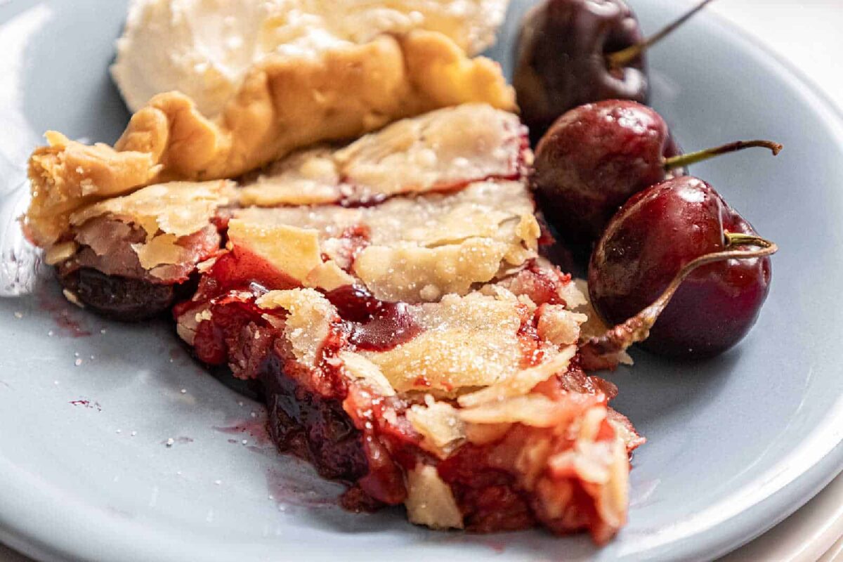 A slice of cherry pie with a flaky crust on a plate, garnished with whole cherries and a piece of pie crust in the background—an irresistible choice for fans of flavorful desserts and classic cherry recipes.