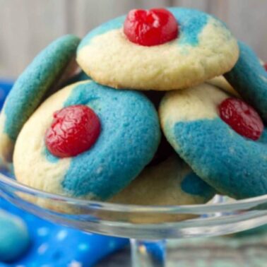 A glass dish holds cookies with blue and beige sections, each topped with a red cherry in the center—perfect for those seeking flavorful desserts or creative cherry recipes.