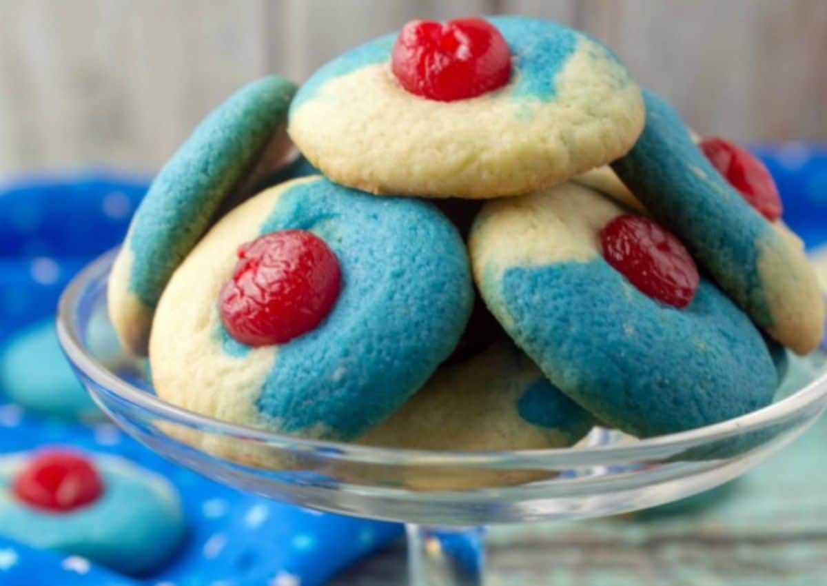 A glass dish holds cookies with blue and beige sections, each topped with a red cherry in the center—perfect for those seeking flavorful desserts or creative cherry recipes.