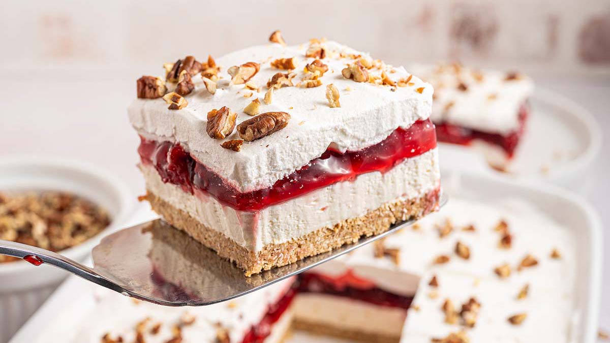 A square slice of layered cherry dessert with a graham cracker crust, creamy filling, cherry topping, whipped cream, and chopped pecans on top, held on a spatula above a baking dish—a perfect addition to your collection of flavorful desserts.
