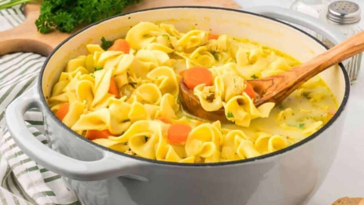 A large pot of comfort food, this soup is filled with wide egg noodles, sliced carrots, herbs, and broth, with a wooden spoon stirring the mixture—perfect for those seeking hearty fall soups or inspiring new soup recipes.
