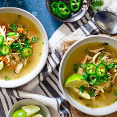 Two bowls of chicken soup with lime wedges, sliced jalapeños, and cilantro on top, set on a striped cloth—this dish brings Main Character Energy to your table. Perfect for anyone looking for flavorful 30 Minute Dinners or quick dinner recipes.