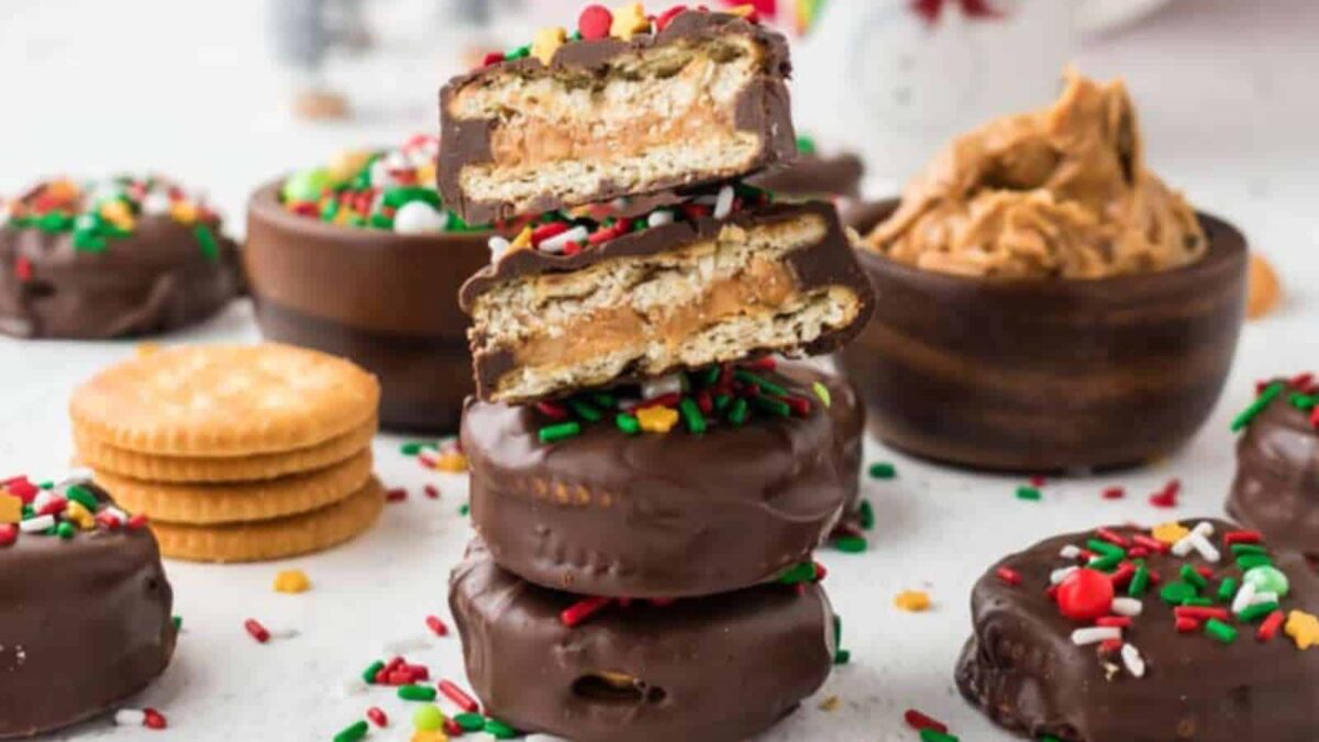 Homemade chocolate treats, these chocolate-covered sandwich cookies filled with peanut butter and topped with colorful holiday sprinkles are stacked with more cookies and peanut butter in the background.