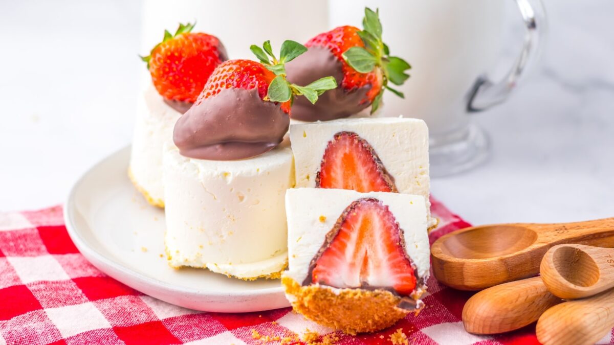 Slices of cheesecake with whole strawberries inside, topped with homemade chocolate treats and chocolate-dipped strawberries, served on a white plate with a red checkered cloth and wooden spoons.