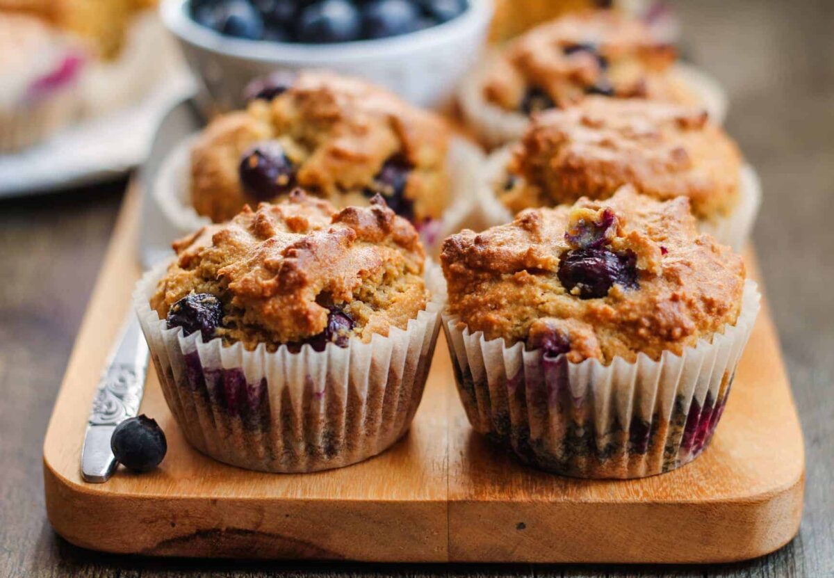 Two blueberry muffins on a wooden board with more muffins and a bowl of blueberries in the background—perfect for anyone looking to try new blueberry breakfast recipes.