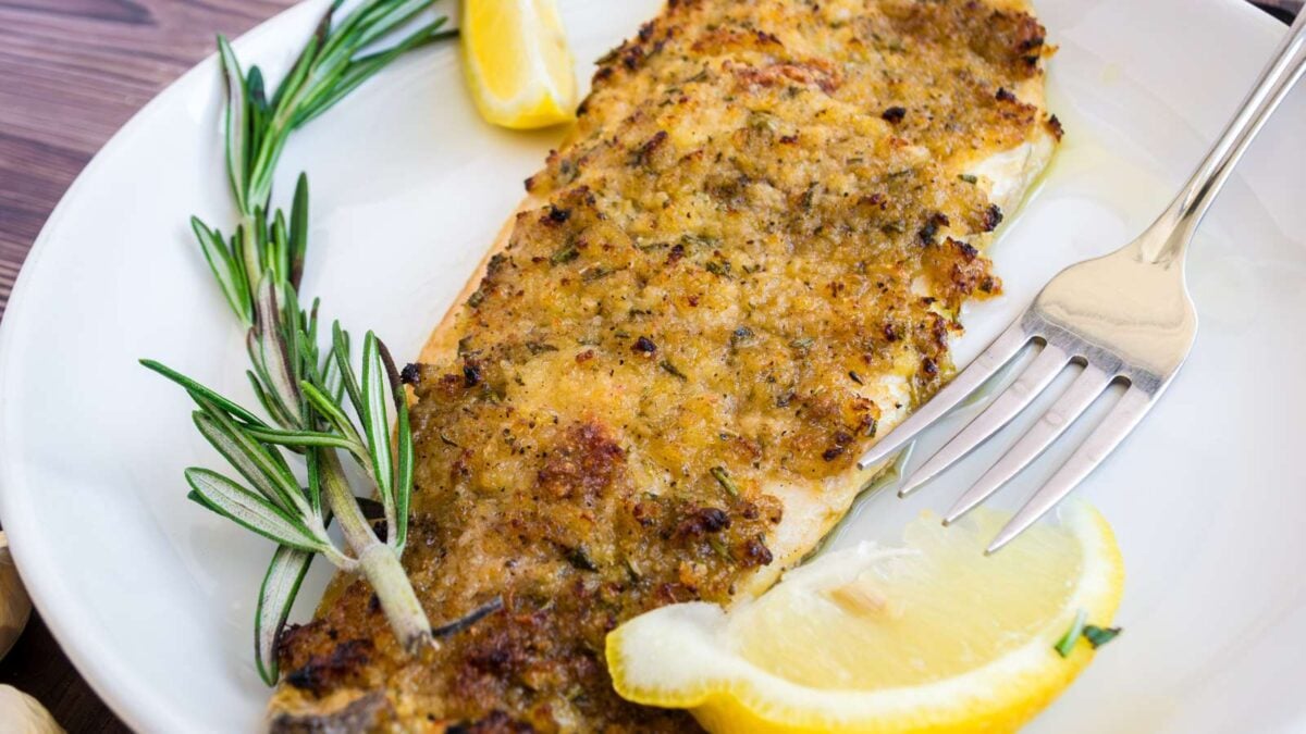 A baked, herb-crusted fish fillet is served on a white plate with a fork, fresh rosemary sprigs, and lemon wedges—perfect for dinner and ideal for fans of simple recipes.