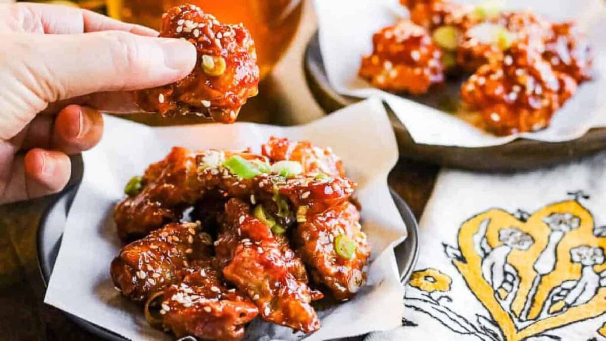 A hand holds a piece of saucy, garnished crispy fried chicken above a plate of similar pieces, with even more tempting chicken recipes displayed on another plate in the background.