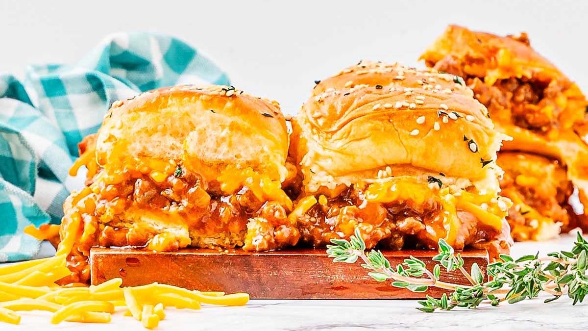 Three sloppy joe sliders with sesame seed buns and melted cheese are arranged on a wooden board, garnished with fresh herbs and shredded cheddar—a perfect choice for easy sliders or new slider ideas. A blue plaid cloth sits in the background.