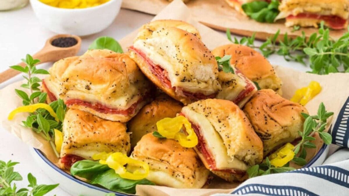 A plate of stacked baked sliders filled with deli meat and melted cheese, garnished with herbs and sliced yellow peppers—perfect for trying new slider recipes or easy sliders at your next gathering.