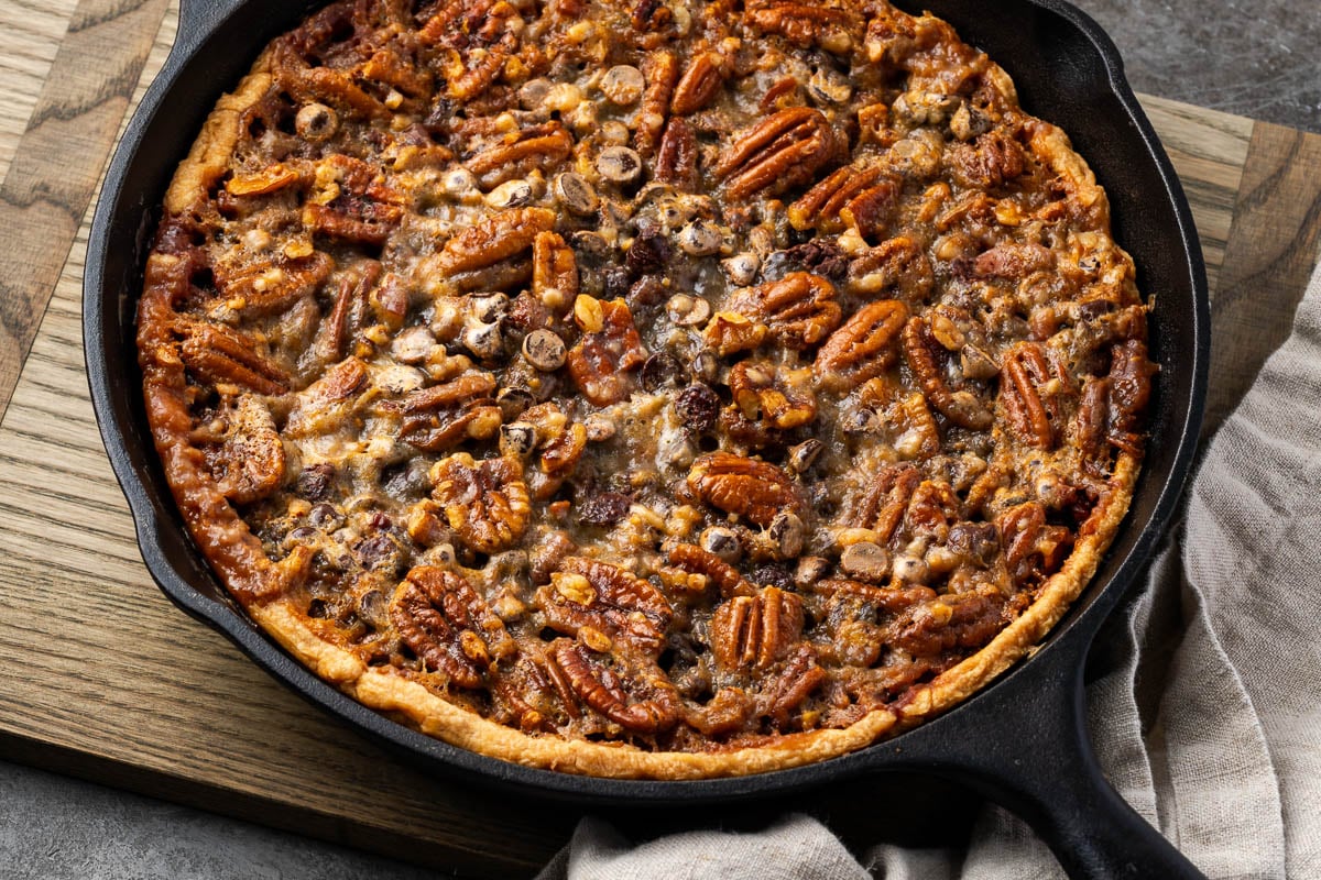 A baked pecan pie in a cast iron skillet sits on a wooden board with a beige cloth napkin underneath, making it a perfect centerpiece for pecan desserts lovers searching for new dessert recipes to try.