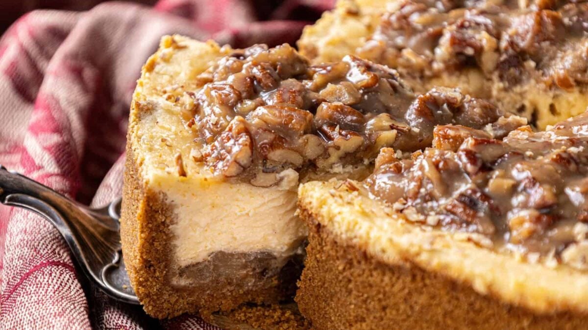 A close-up of a sliced pecan cheesecake with a crumbly graham cracker crust, creamy filling, and pecan topping on a red-patterned cloth—perfect for fans of pecan desserts seeking new dessert ideas.