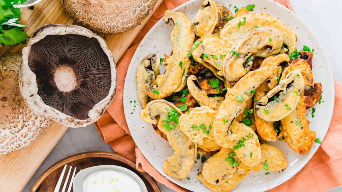 A plate of battered and fried mushroom slices, a creative fries twist, garnished with chopped parsley, sits next to whole portobello mushrooms and a small bowl of dipping sauce.