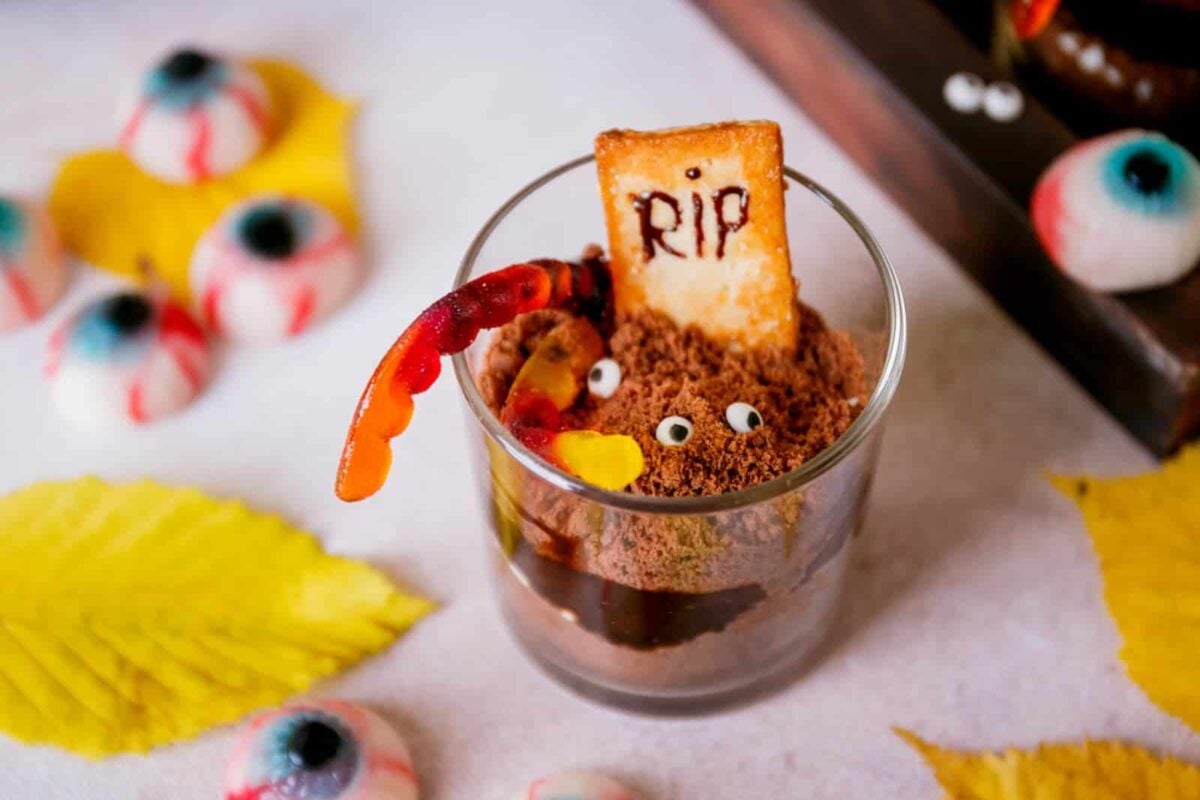 A glass of chocolate dessert decorated with candy eyes, a gummy worm, and a cookie marked "RIP," surrounded by fake eyeballs and yellow leaves—perfect for fans of Monster Sweets or Monster Themed Halloween Sweets.