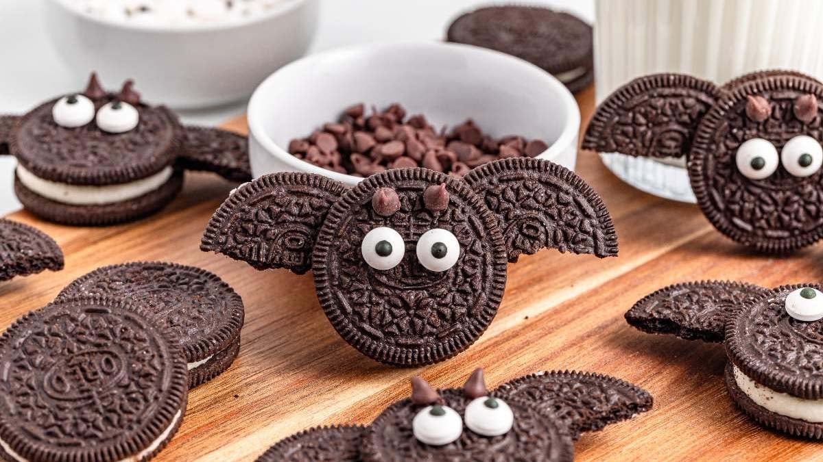 Chocolate sandwich cookies decorated with candy eyes and extra cookie wings to resemble bats make spooky Monster Themed treats, arranged on a wooden board near a bowl of chocolate chips and a glass of milk.