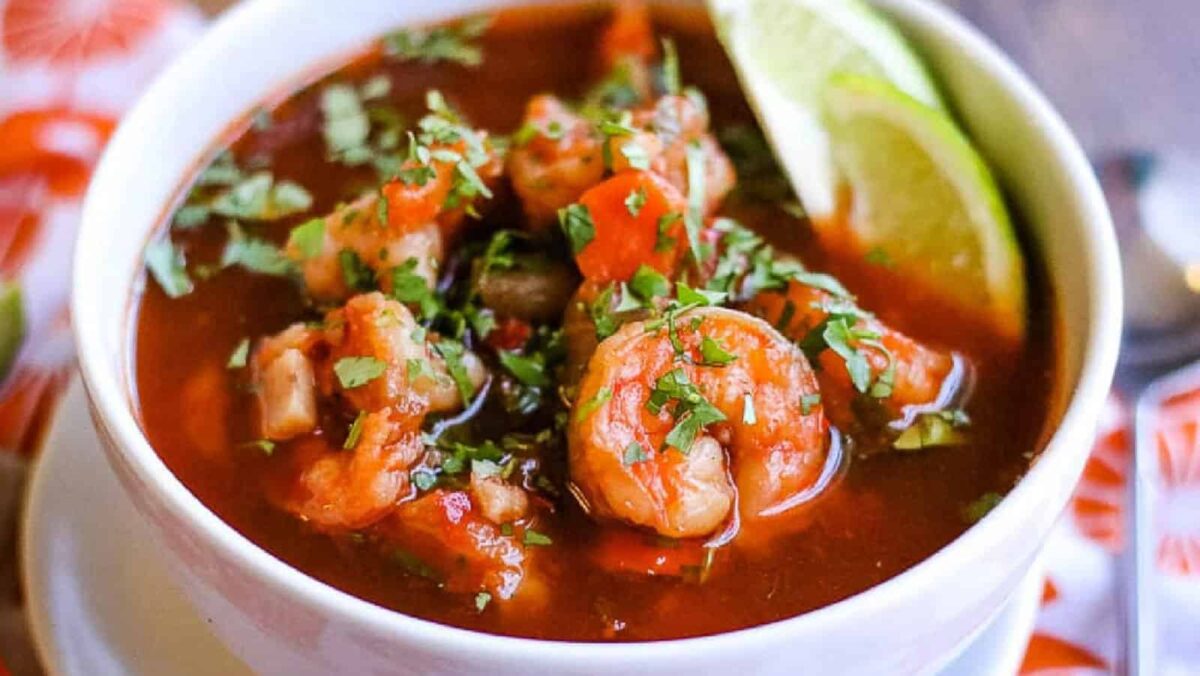 A white bowl filled with shrimp soup, garnished with chopped cilantro and lime wedges, sits on a table—perfect for easy dinners or quick dinner recipes.