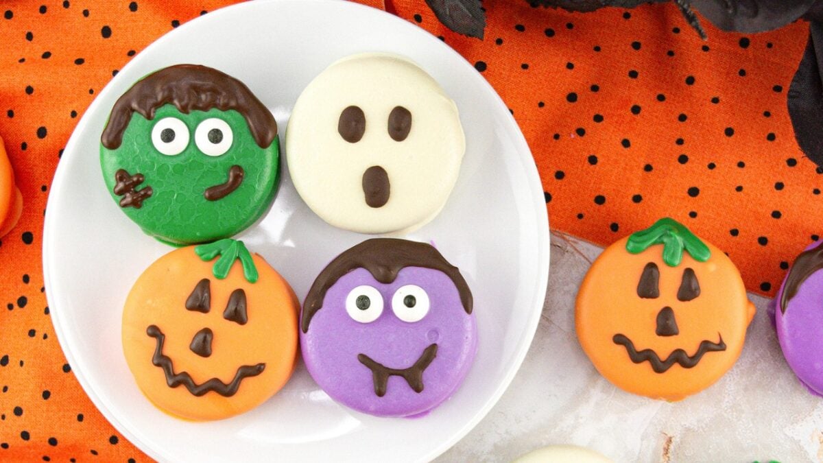 A plate of Monster Themed Halloween Sweets featuring a green monster, a white ghost, a purple monster, and two orange pumpkins on an orange polka dot tablecloth.