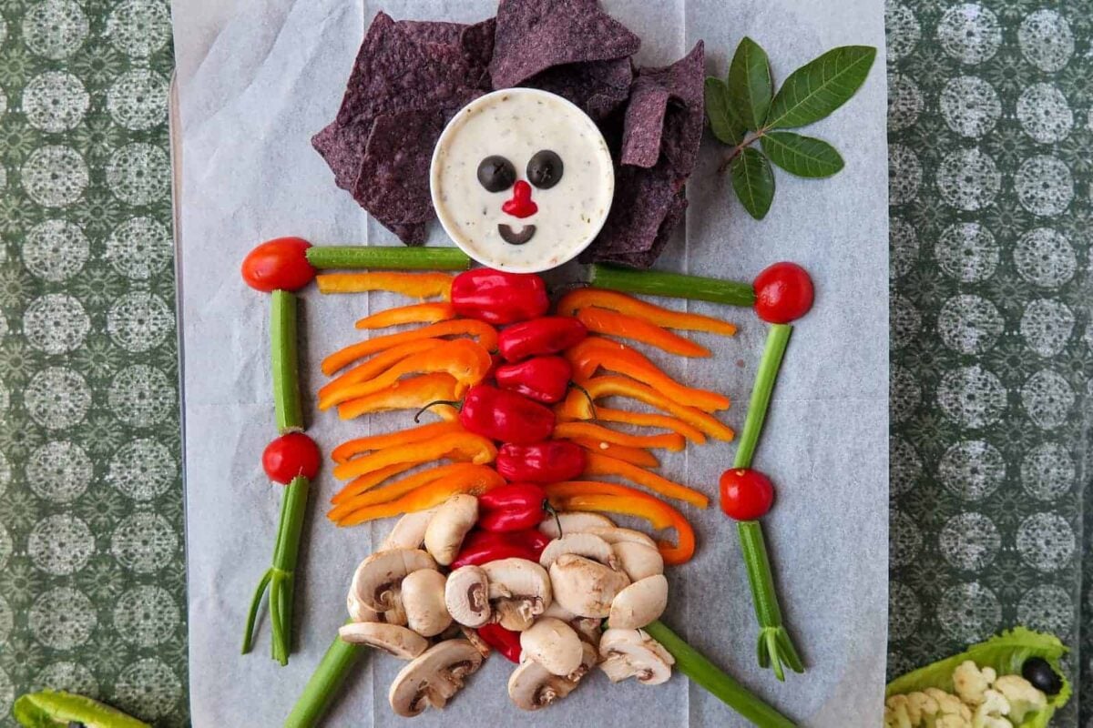 Vegetables and chips are arranged on a tray to resemble a skeleton, making it a fun Halloween appetizer—perfect for any Halloween party. Dip and olives form the head, with greens, peppers, tomatoes, and mushrooms creating the body and limbs.