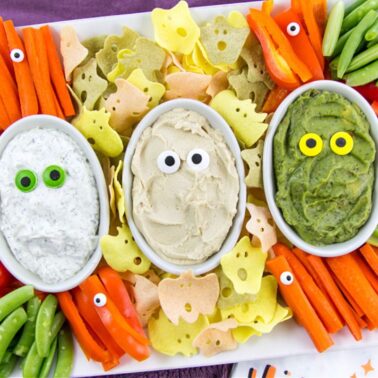 A festive vegetable platter with three dips in oval bowls, each decorated with candy eyes, surrounded by sliced carrots, celery, bell peppers, snap peas, and Halloween-themed chips—perfect for Halloween appetizers at your next party.