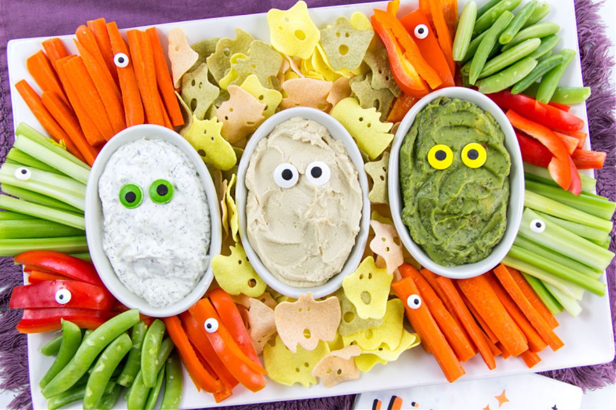 A festive vegetable platter with three dips in oval bowls, each decorated with candy eyes, surrounded by sliced carrots, celery, bell peppers, snap peas, and Halloween-themed chips—perfect for Halloween appetizers at your next party.