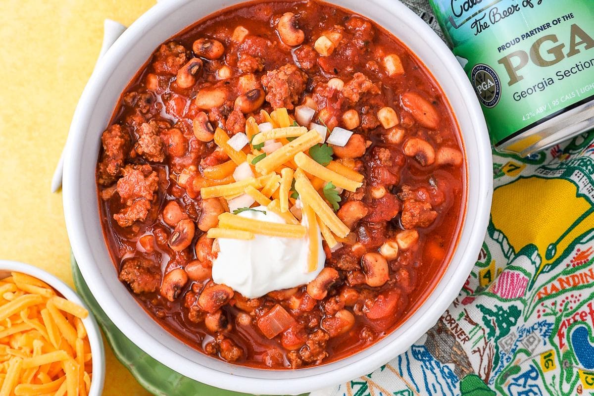 A bowl of easy chili with ground meat, beans, topped with shredded cheese, sour cream, and chopped onions—perfect for lazy weeknight dinners—sits next to a can and a bowl of extra cheese.