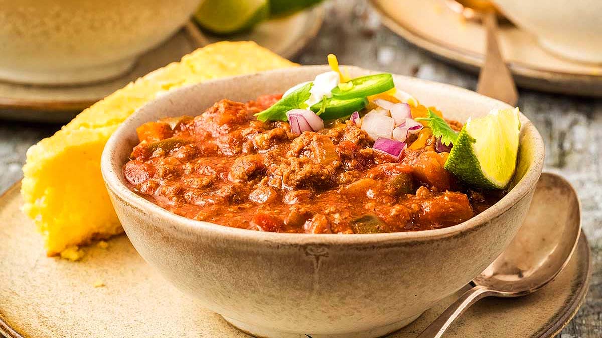 A bowl of easy chili topped with cheese, onions, and a lime wedge, served with a slice of cornbread on a plate with a spoon—perfect for cozy weeknight dinners.