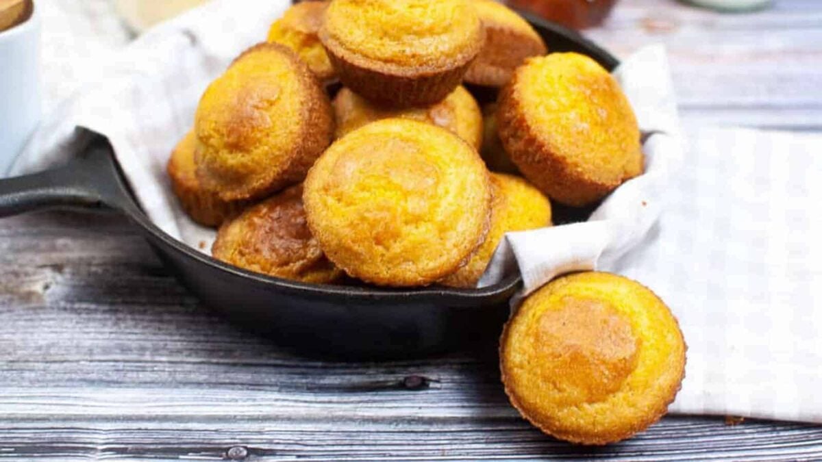 A cast iron skillet lined with a white towel holds several golden brown cornbread muffins, comfort food at its best, with one muffin resting on the wooden surface nearby—perfect for fans of easy baking or classic cornbread recipes.