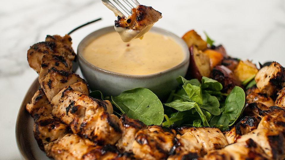 Grilled chicken skewers arranged around a bowl of sweet and savory dipping sauce, with spinach leaves and roasted vegetables on the side. A fork dips a chicken piece into the sauce—perfect for those who love to cook with apple cider.
