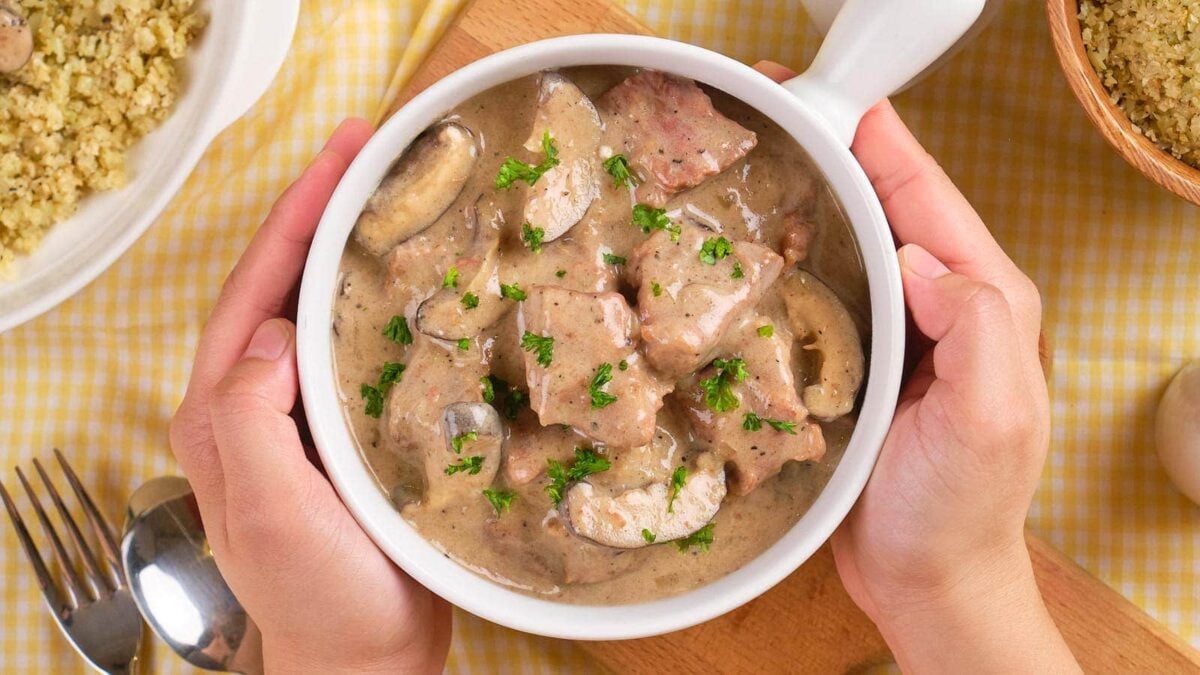 A bowl of easy beef stroganoff with mushrooms and creamy sauce, garnished with chopped parsley, held by two hands over a yellow checkered tablecloth—a perfect pick for dinner or anyone who loves slow cooker beef recipes.