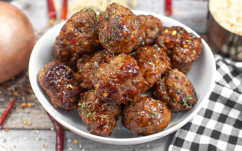 A white bowl filled with glazed, browned meatballs, garnished with herbs, sits on a wooden surface next to a black and white checkered cloth—perfect for easy beef recipes and quick dinner ideas.