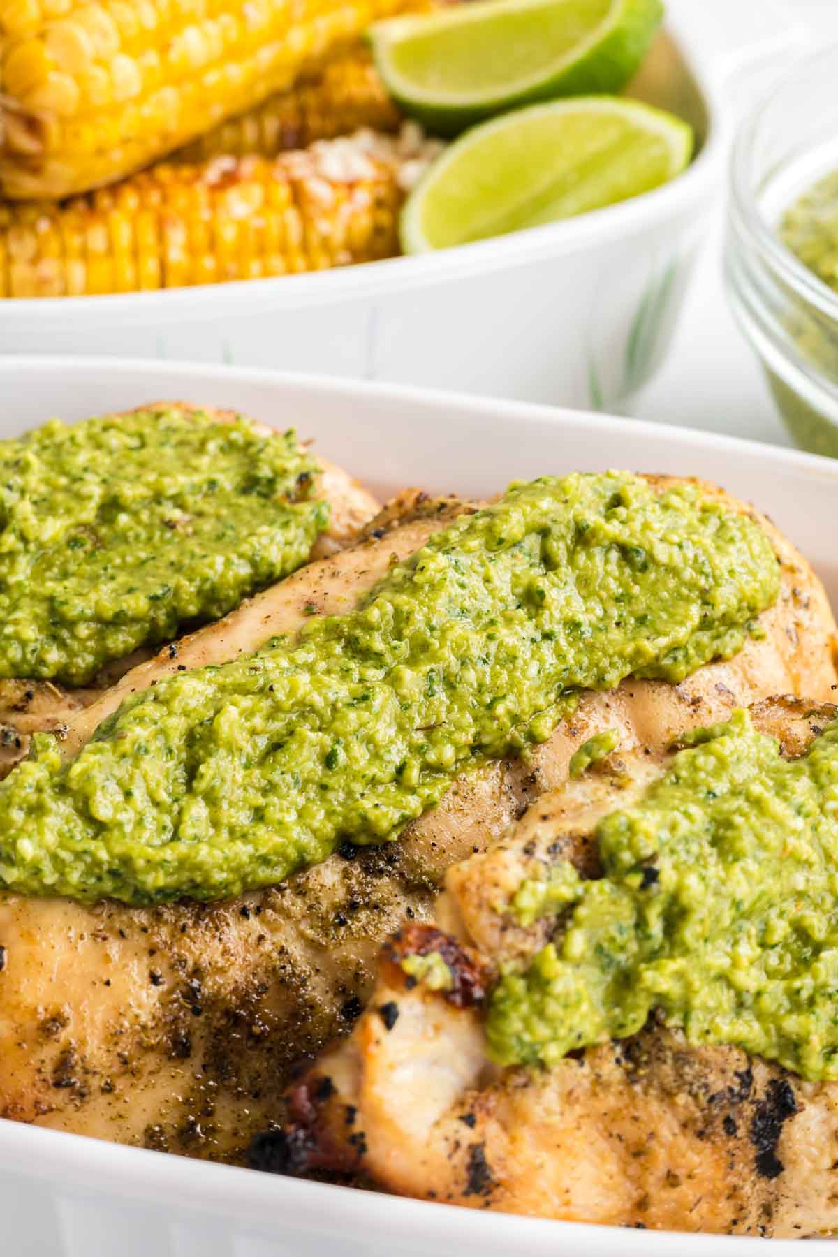 Grilled chicken breasts topped with jalapeno pesto in a white dish, with corn on the cob and lime wedges in the background.