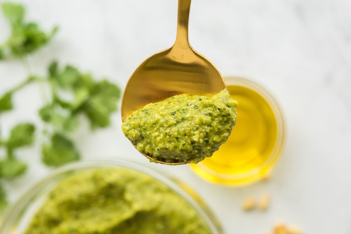 A gold spoon holds a portion of roasted jalapeno pesto sauce above a bowl, with olive oil and cilantro visible in the background.