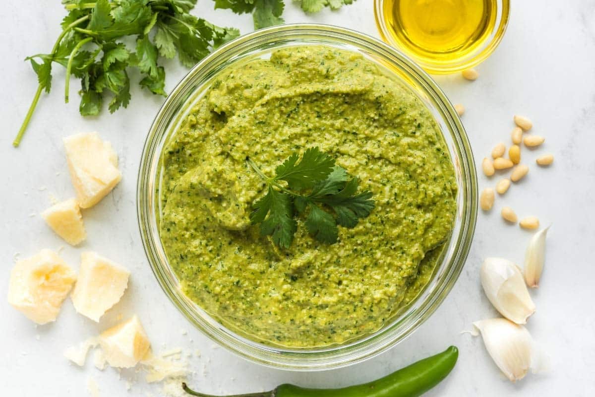 A bowl of jalapeno pesto garnished with cilantro, surrounded by parmesan cheese, cilantro, olive oil, pine nuts, garlic cloves, and a green chili pepper on a white surface.