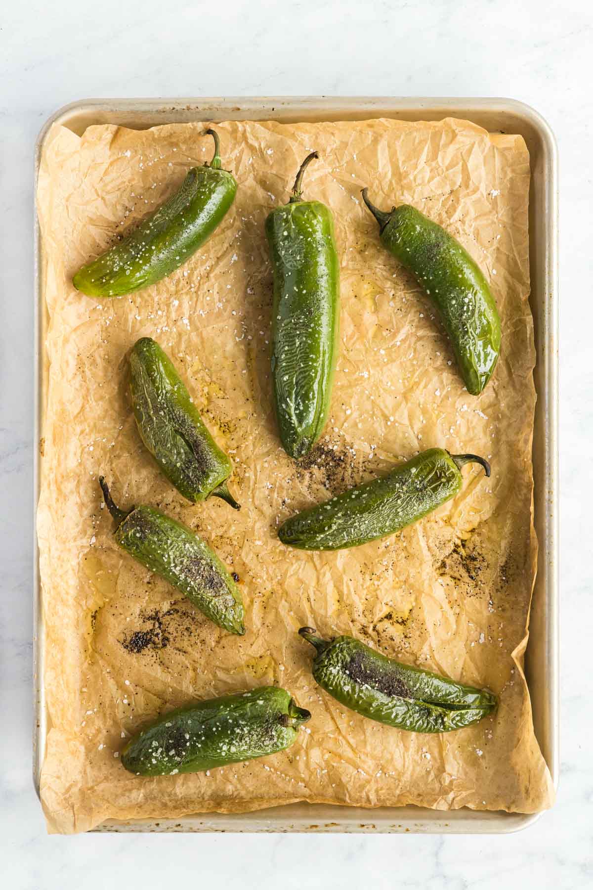 A baking sheet lined with parchment paper holds eight roasted green poblano peppers sprinkled with salt.